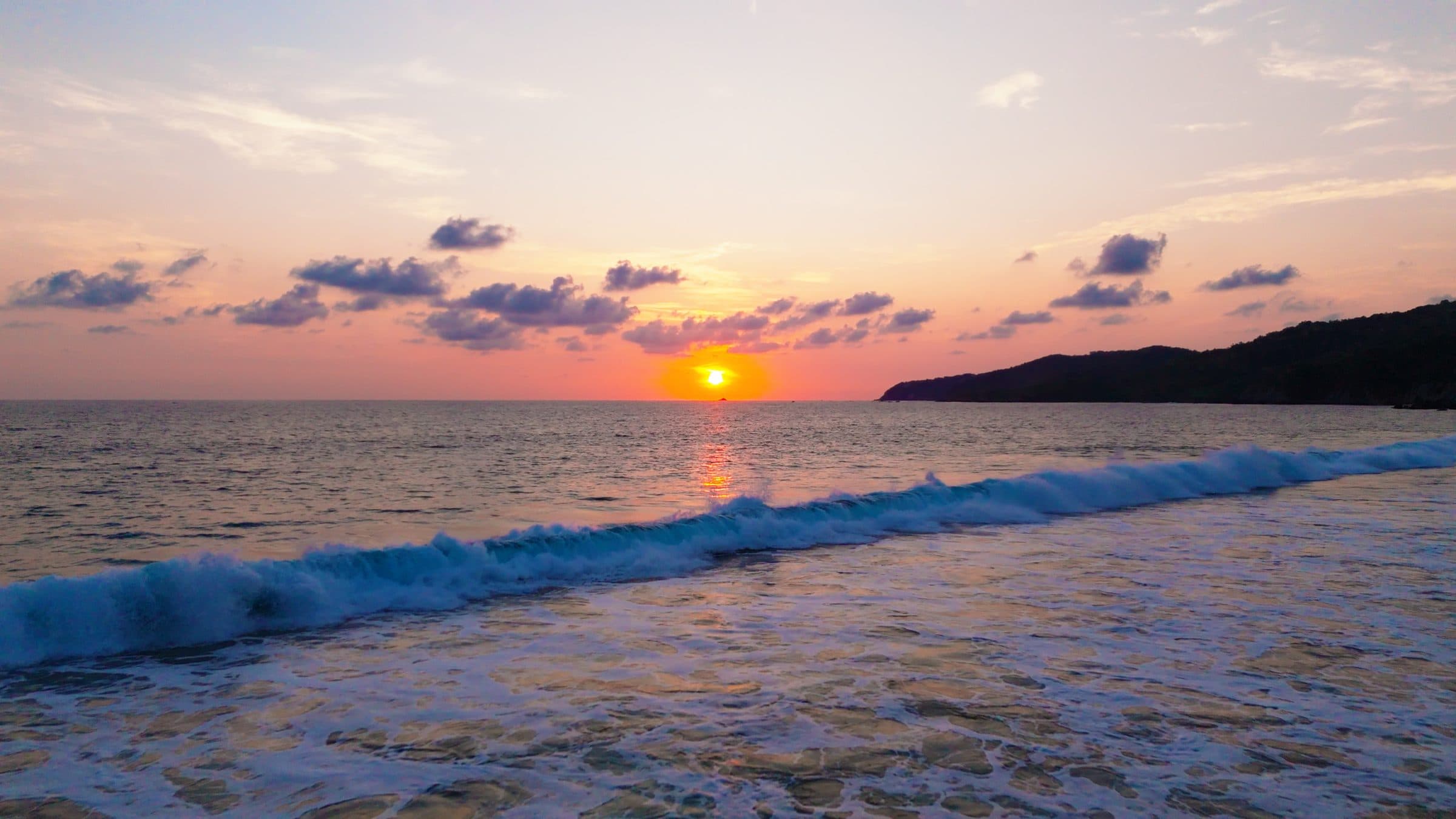 The sun setting into the sea, with a wave breaking on the shore